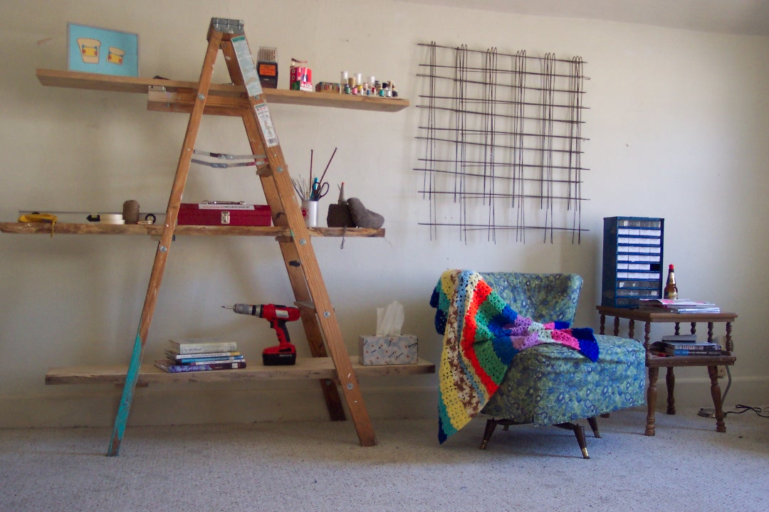 A ladder shelf, wire hangings on a wall and a short arm chair with a quilted throw. 