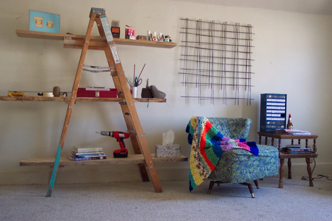 A ladder shelf, wire hangings on a wall and a short arm chair with a quilted throw.