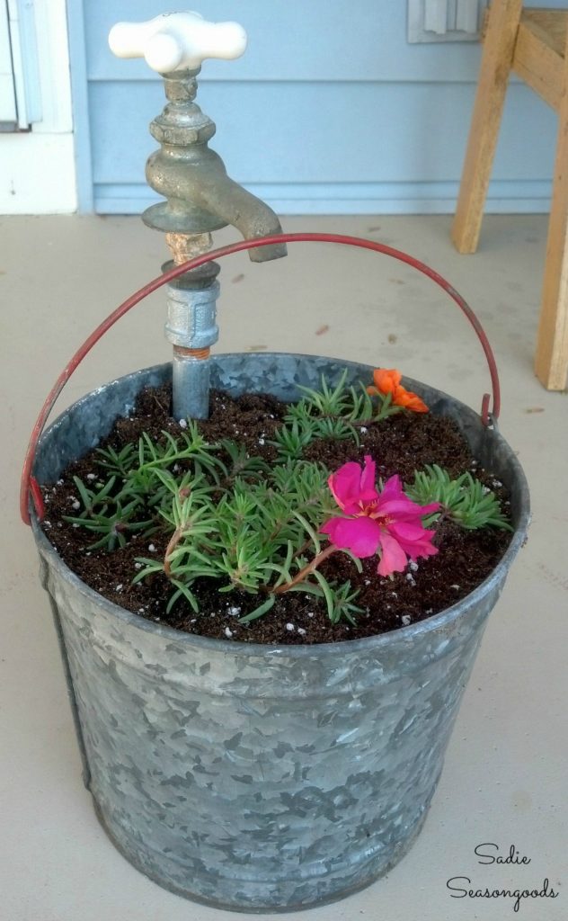 A tin bucket with a faucet attached is filled with soil and plants.